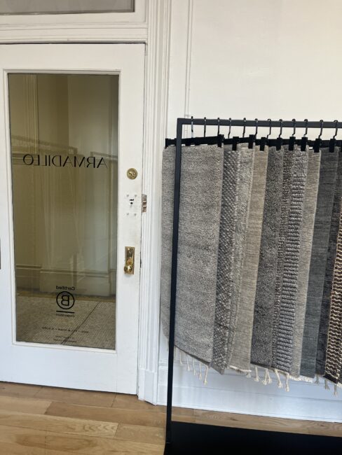A metal rack displays several folded rugs in various gray patterns beside a glass door labeled “ARMADILLO.” The floor is wooden and the walls are white.