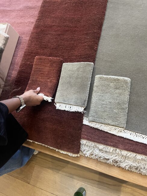 A person holds a red carpet swatch next to two gray swatches, comparing them against a large rug with red and gray sections on a wooden floor.