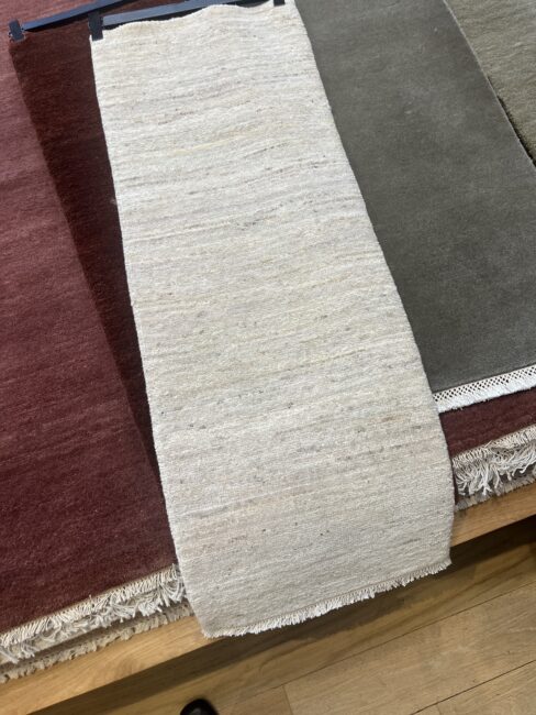 A cream-colored runner rug displayed over larger olive green and burgundy rugs on a wooden surface.