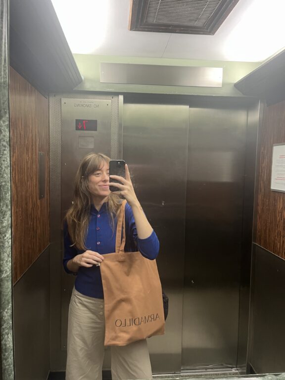 A person stands in an elevator taking a mirror selfie, holding a tan tote bag with the word "ARMADILLO" printed on it.