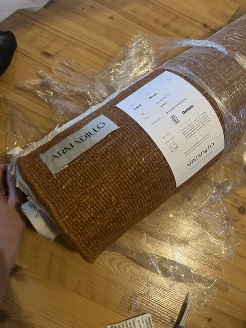A hand unwraps a rolled brown Armadillo rug on a wooden floor, with plastic packaging and product label visible.
