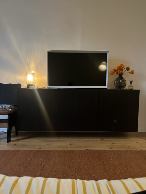 A flat-screen TV on a dark cabinet with a lamp, a book, and a vase of orange flowers in a living room setting.