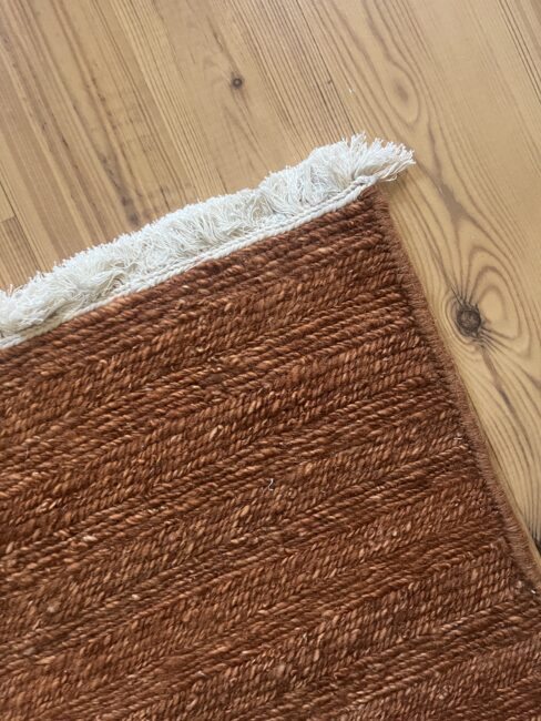 A close-up of a brown woven rug with white fringe, placed on a light-colored wooden floor.