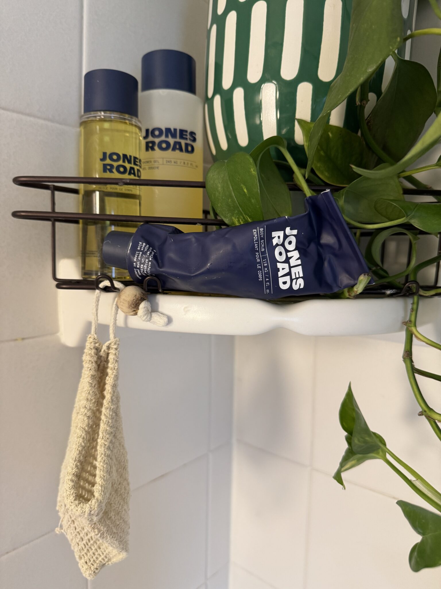 Three Jones Road skincare products and a washcloth sit on a wire bathroom shelf next to an Ilia product and a green leafy plant in a patterned pot, all set against a white tiled wall.
