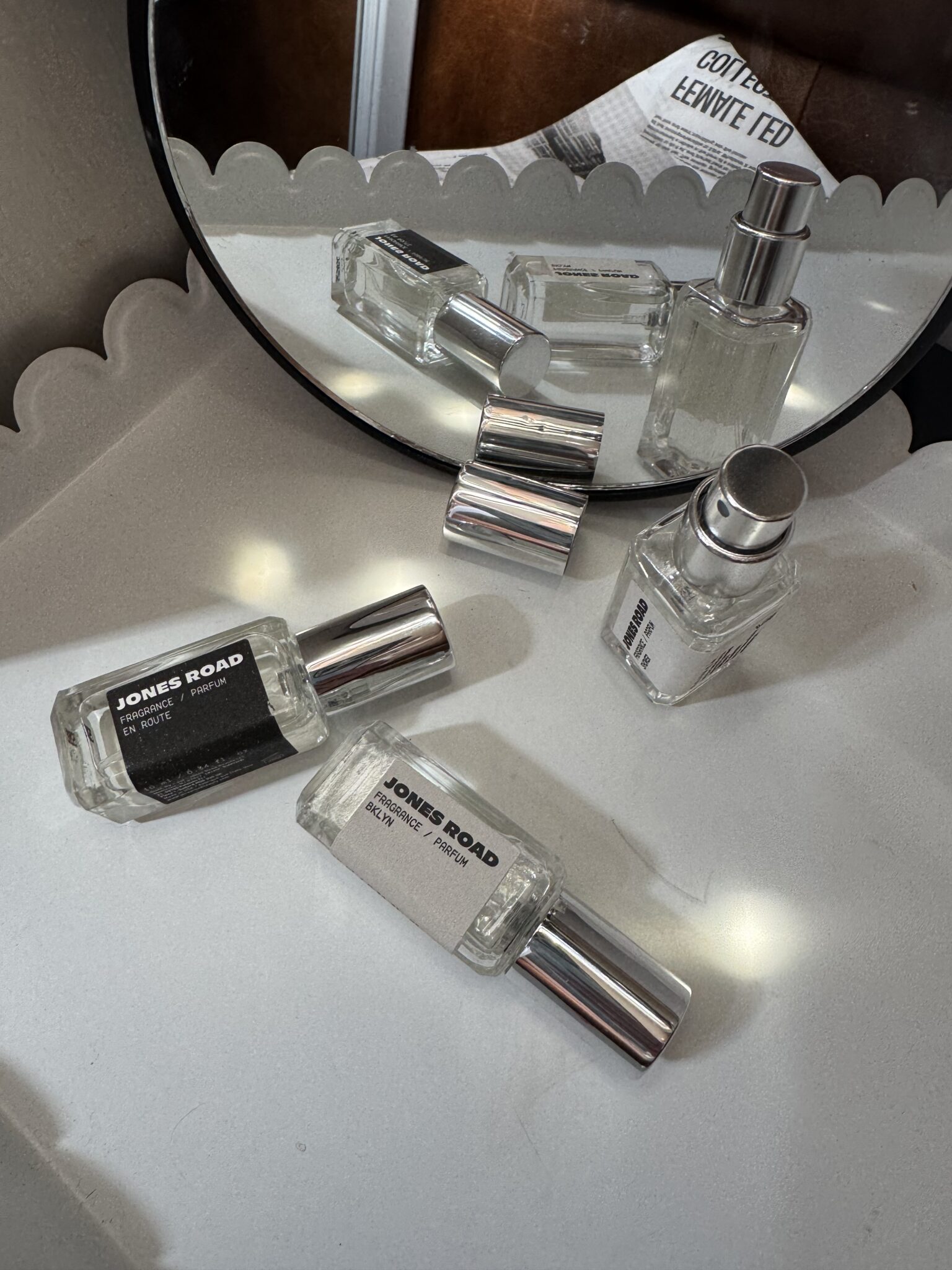 Three Jones Road perfume bottles with silver caps are arranged on a white surface, while two are reflected in a round mirror. A folded newspaper and an Ilia product peek out in the background, creating a chic and modern scene.