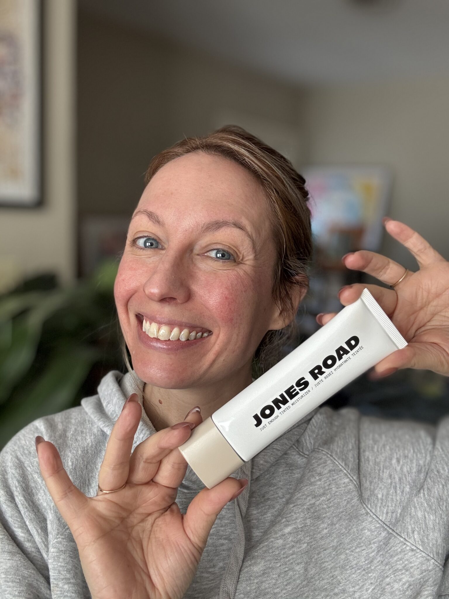 A smiling person in a gray hoodie holds up a tube labeled "JONES ROAD"—a moment reminiscent of Ilia’s minimalist beauty—in front of a neutral indoor background.