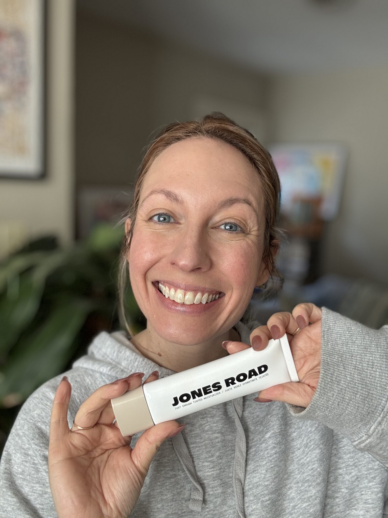 Woman smiling and holding a Jones Road beauty product in a tube, indoors, with plants and artwork in the background—her Ilia favorites nearby suggest she loves clean beauty brands.