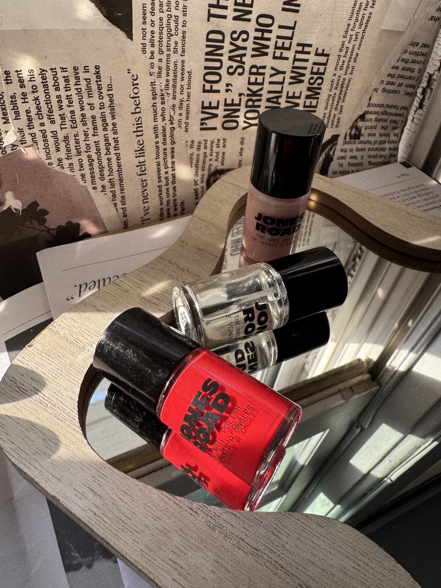 Three bottles of nail polish, including a red Ilia shade, sit on a wooden-framed mirror. Newspapers and papers are scattered in the background.