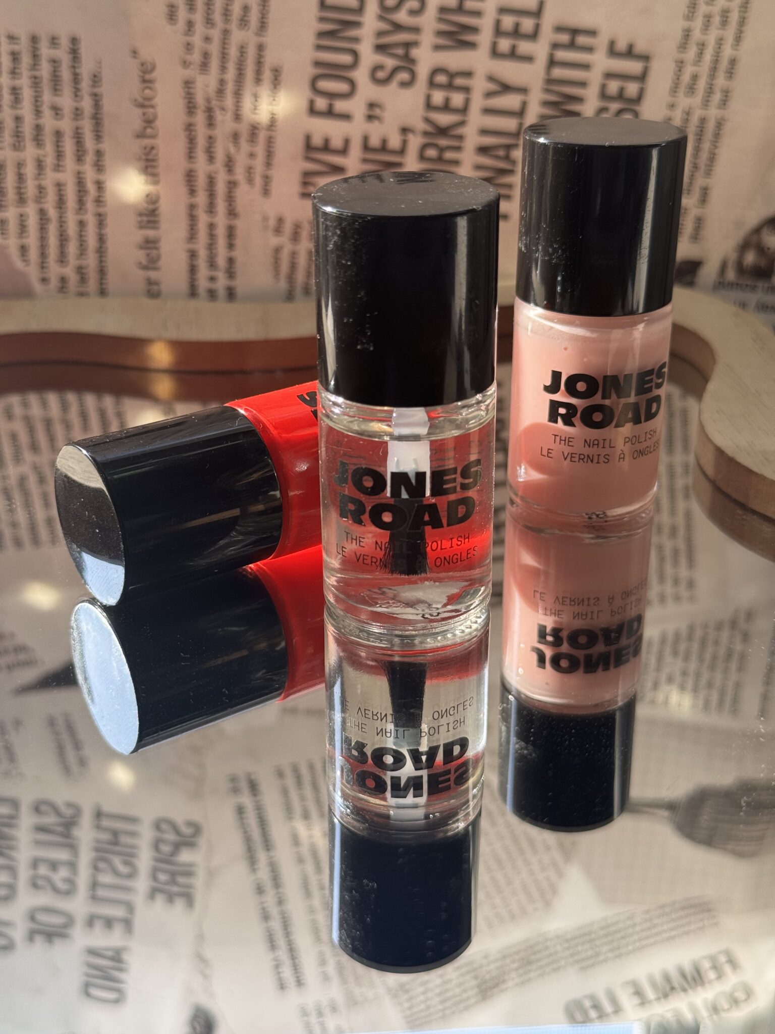 Three Jones Road nail polish bottles—one clear, one red, and one light pink—are arranged on a reflective surface with a newspaper print background, creating a chic look that would pair seamlessly with any Ilia beauty collection.