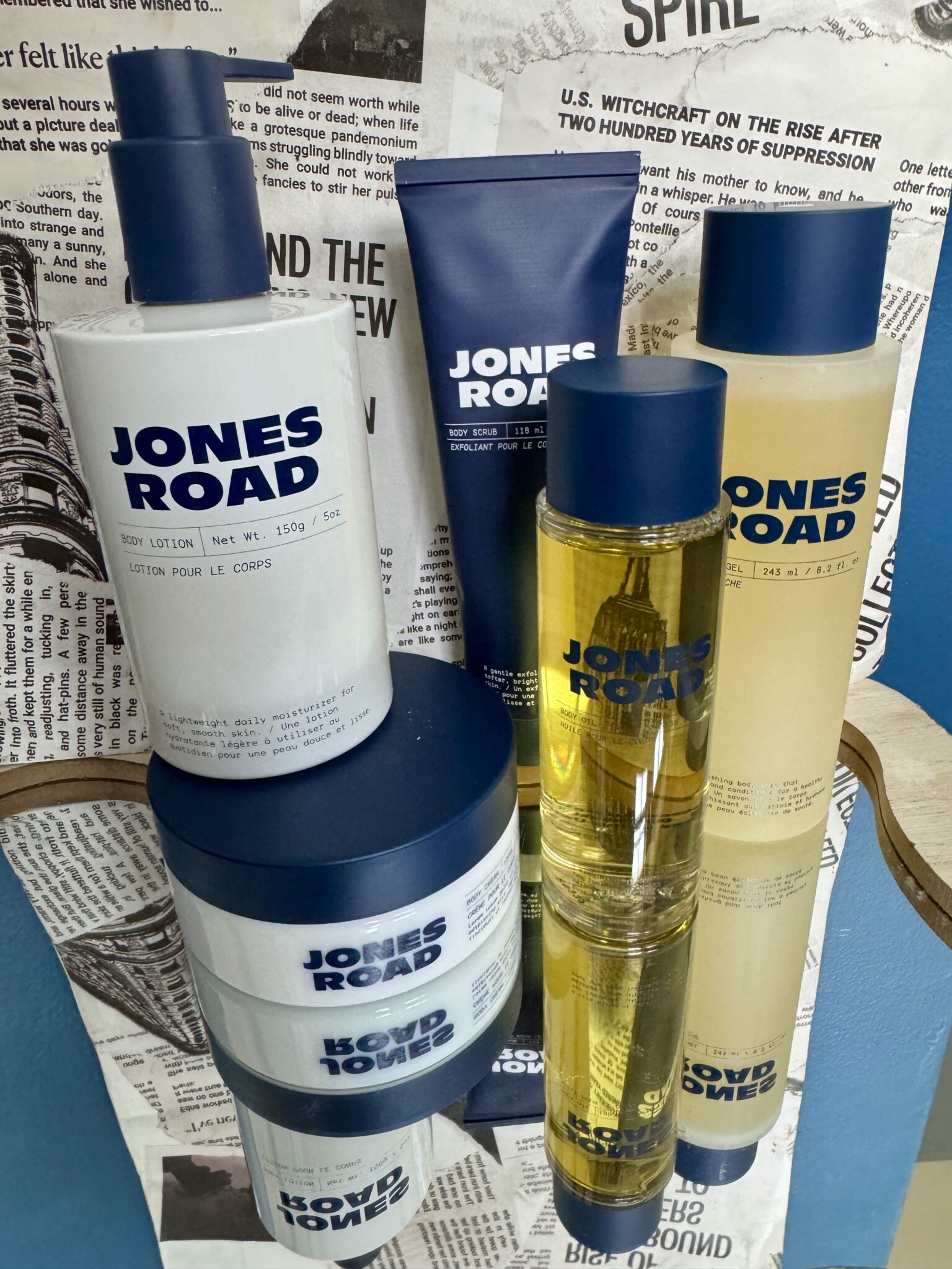 Several Jones Road and Ilia skincare products, including bottles, jars, and tubes, are arranged in front of a collage of newspaper clippings on a wall.