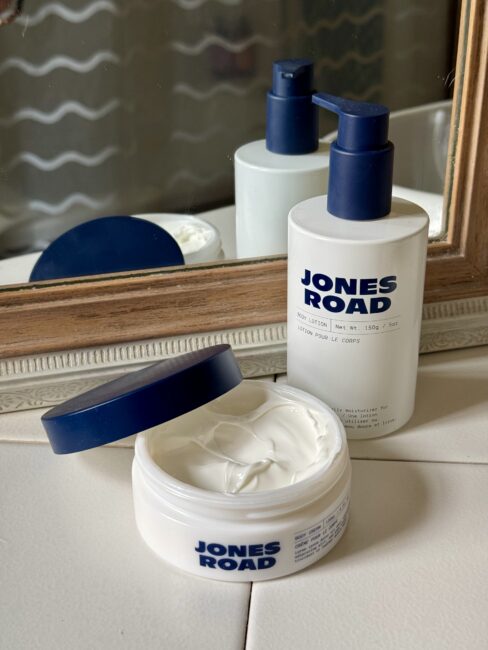 Two Jones Road skincare products, a body lotion with a pump and an open jar of cream, are placed on a bathroom counter in front of a mirror, creating a serene scene alongside Ilia favorites.