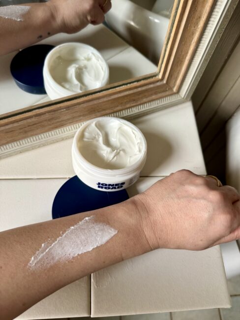 A hand with a streak of white Ilia cream applied on the forearm is shown near an open container of cream, placed on a tiled bathroom counter beside a mirror.