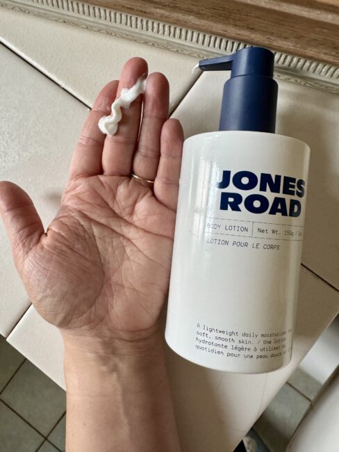A hand holding a small amount of white lotion next to a bottle labeled "Jones Road Body Lotion" on a tiled surface, reminiscent of the clean aesthetic seen in Ilia skincare imagery.