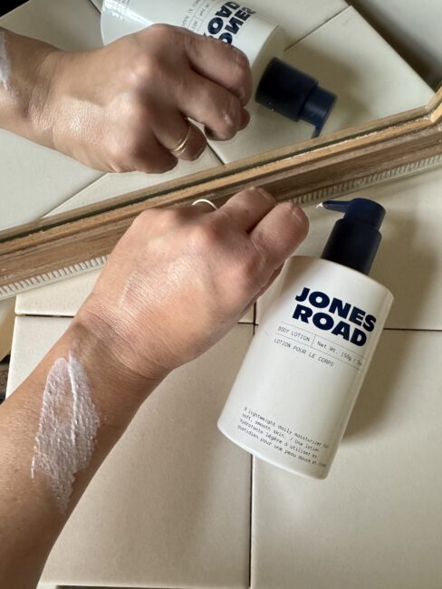A hand with lotion applied is shown next to a bottle of Jones Road Body Lotion and an Ilia product, with both reflected in a mirror.