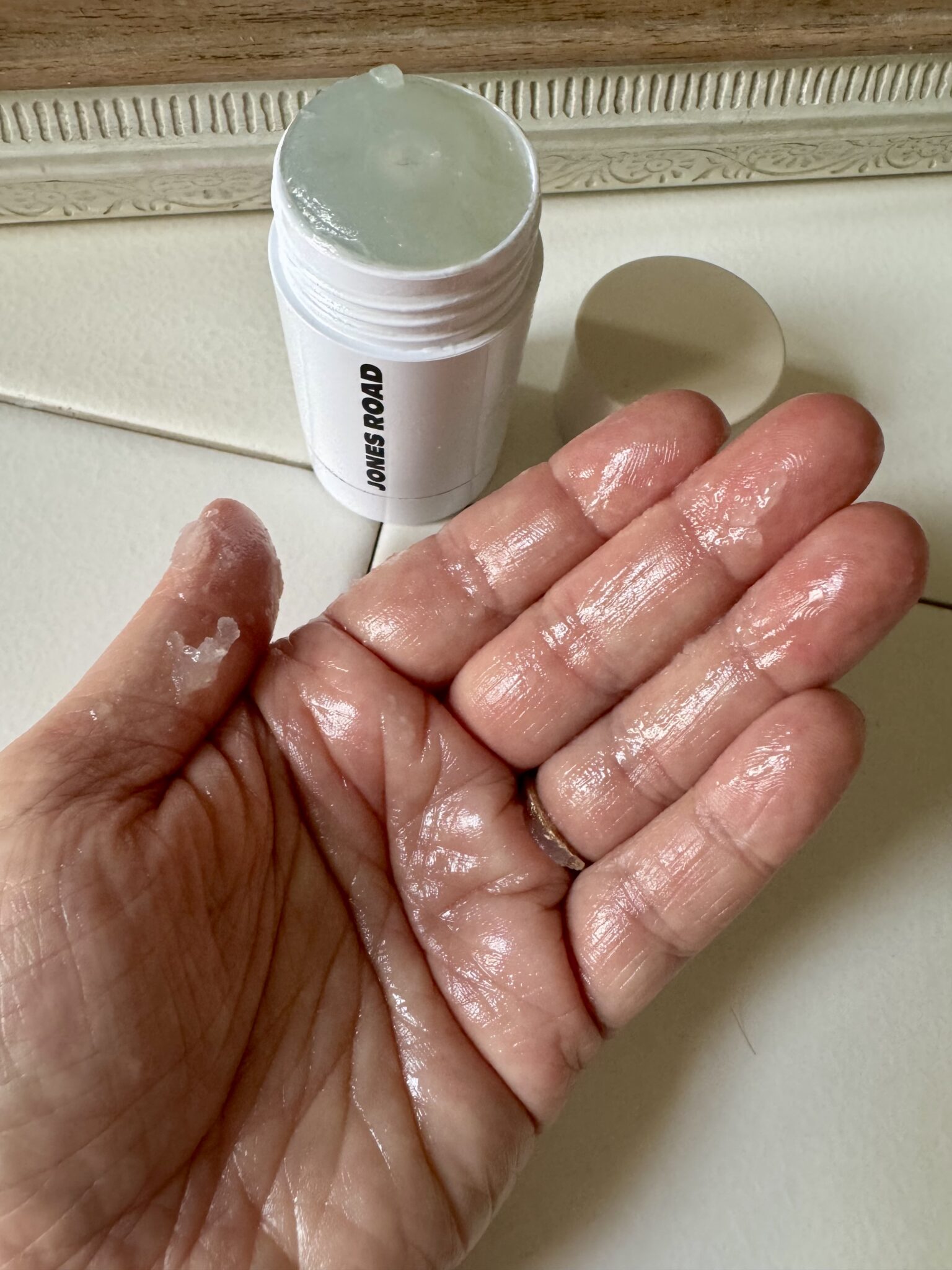 A hand with a shiny substance on the palm is shown in front of an open stick container labeled "JONES ROAD," reminiscent of popular brands like Ilia.