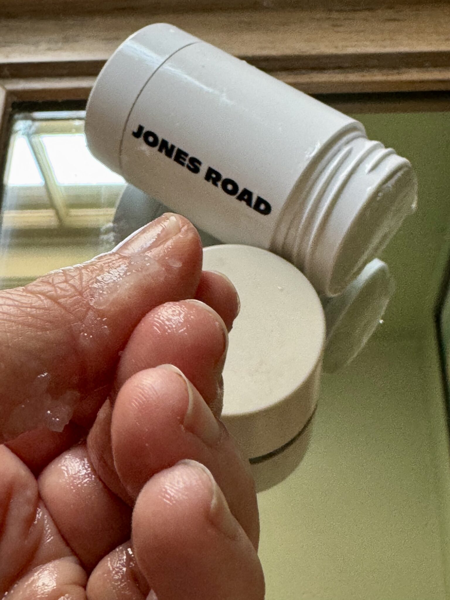 A hand with damp fingers holds a small amount of product near an open Jones Road stick container and an Ilia item on a mirrored surface.