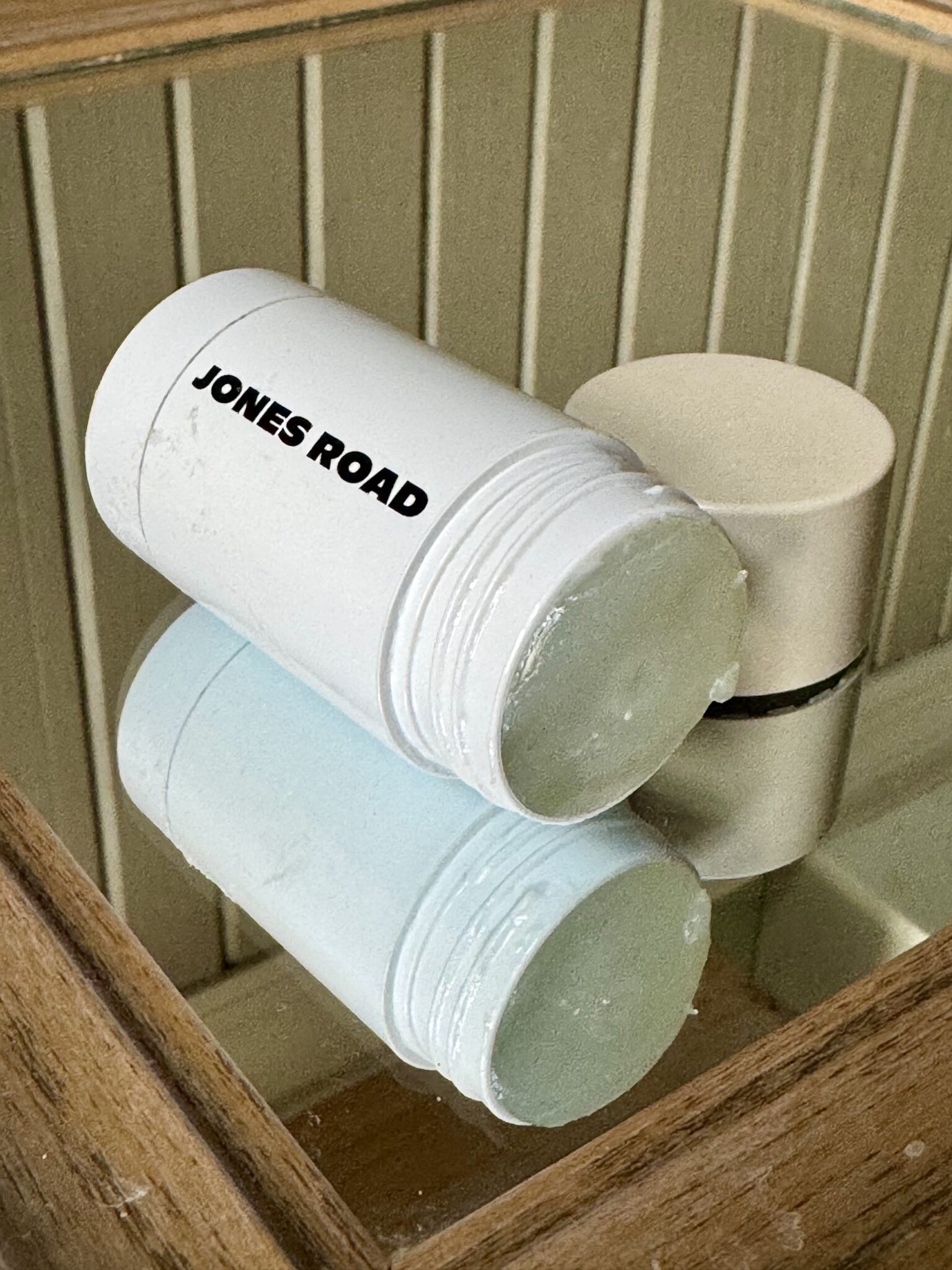 A Jones Road balm stick with the cap off lies on its side on a mirrored surface, reflecting the product and surrounding wooden frame—a chic scene reminiscent of Ilia’s minimalist beauty aesthetic.