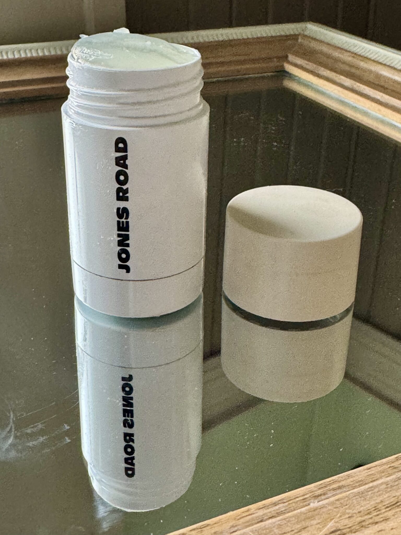 A white Jones Road stick container with its cap off sits on a mirrored surface, reflecting the product, its label, and offering a sleek look reminiscent of Ilia’s minimalist design.