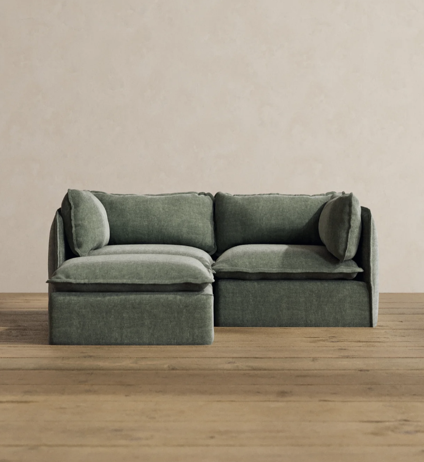A green sectional sofa with two seat cushions, two back cushions, and two side pillows on a wooden floor against a beige wall.