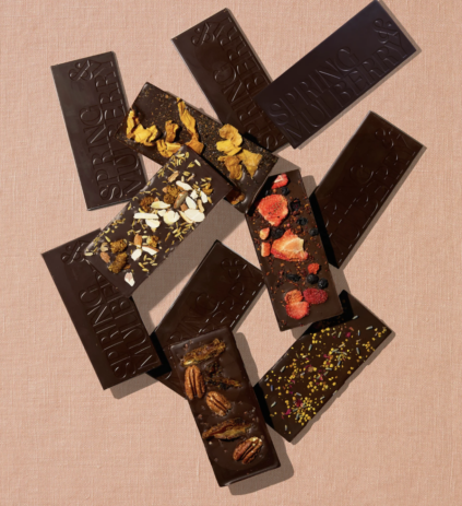 Assorted dark chocolate bars, some topped with nuts, dried fruit, cornflakes, or sprinkles, displayed on a pink textured surface.