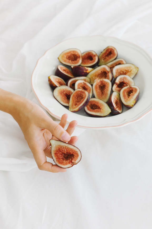 A hand holds a halved fig above a white plate filled with more halved figs, all set on a white fabric background.