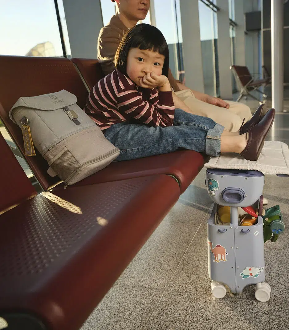 A young child sits on an airport bench with feet up on a small suitcase decorated with animal stickers; an adult sits nearby and a backpack rests on the seat.