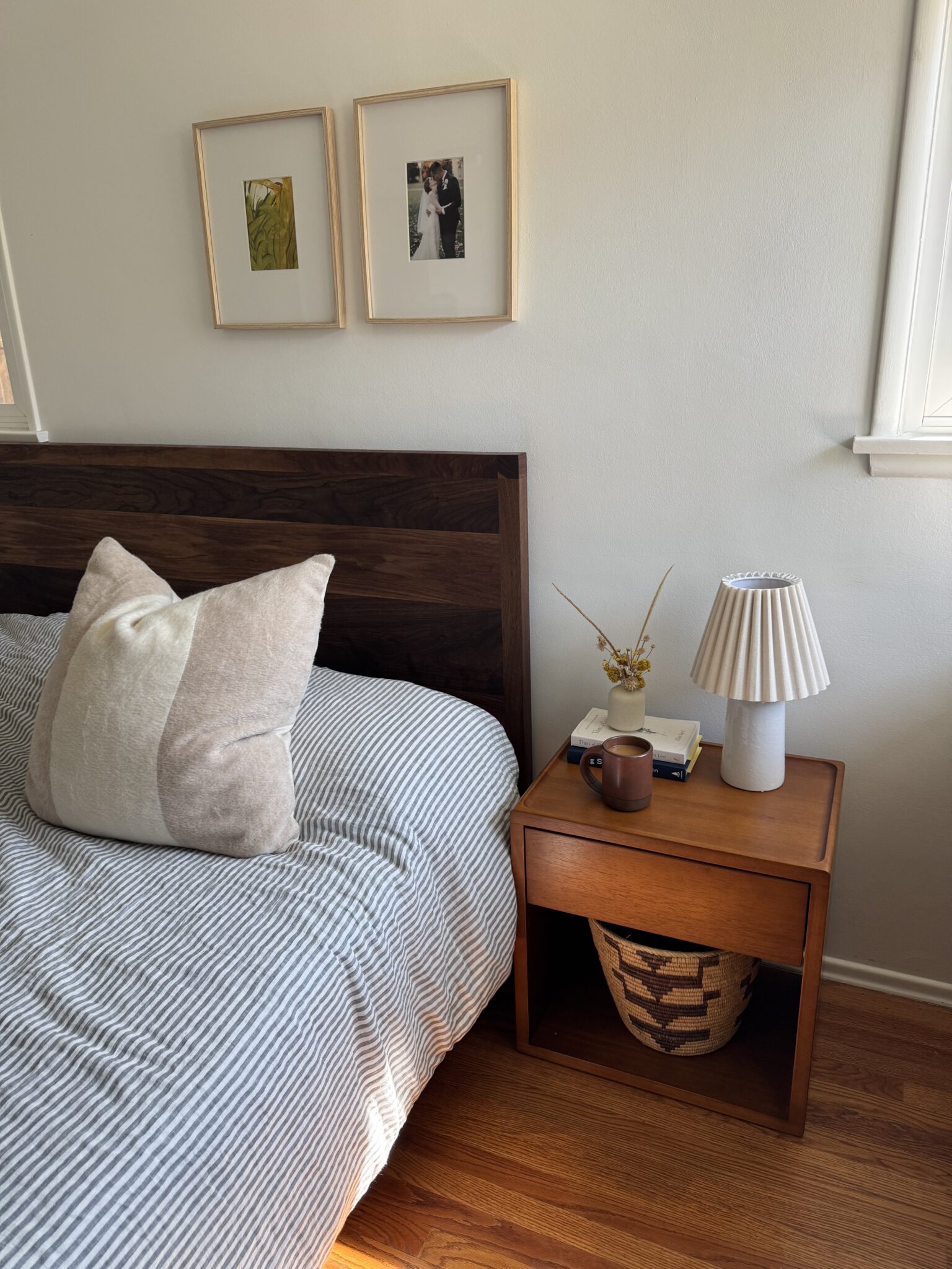 A neatly made bed with a striped duvet, a large pillow, a wooden headboard, and a nightstand holding a lamp, mug, books, and dried flowers, with framed photos on the wall above.