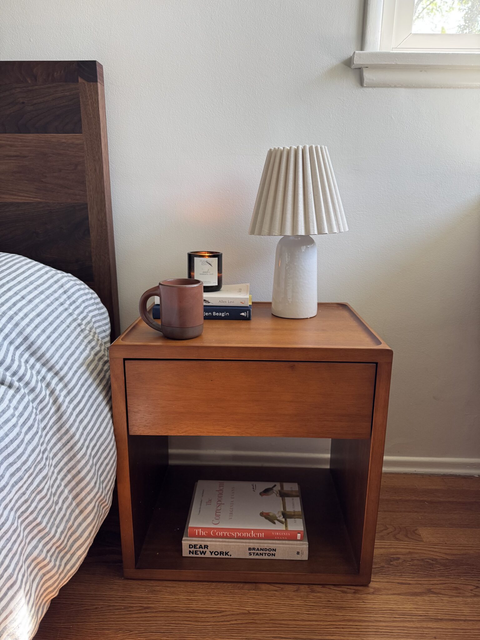 A wooden nightstand with a small lamp, a mug, a candle, and books on top, with more books on the lower shelf, next to a bed and under a window.