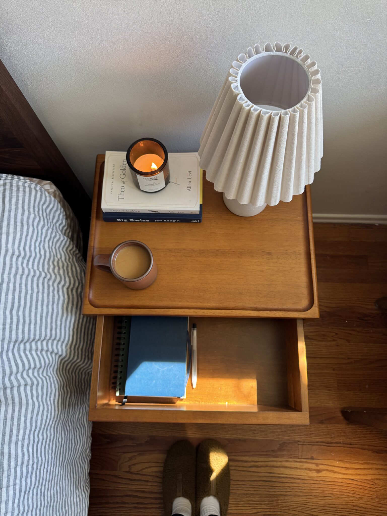 A wooden bedside table with a lit candle, a stack of books, a cup of coffee, a closed notebook, a lamp, and an open drawer; slippers and feet are visible at the bottom.