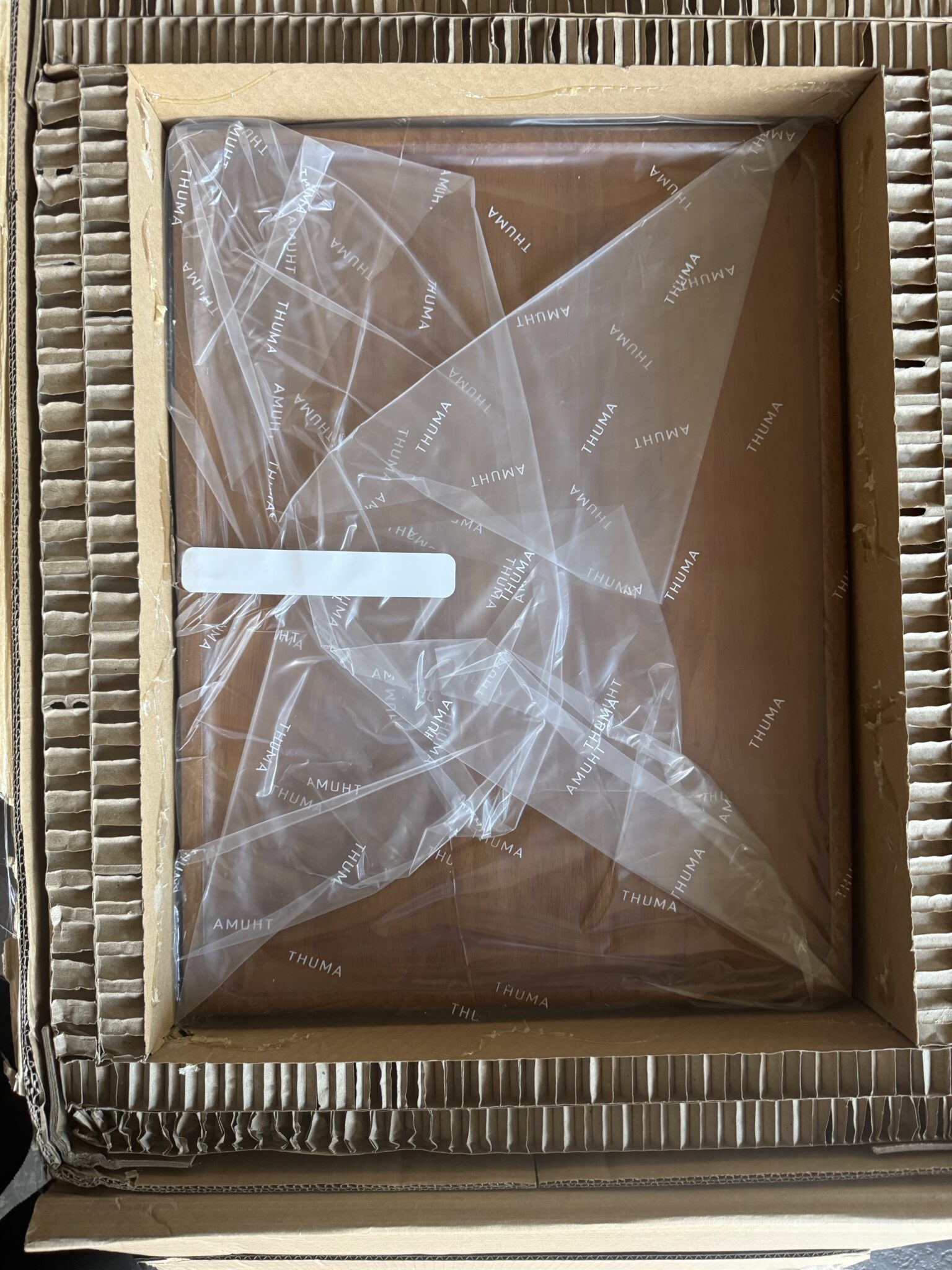 A wooden panel labeled "THUMA" is packaged inside a cardboard box with protective corrugated paper and covered in plastic wrapping.