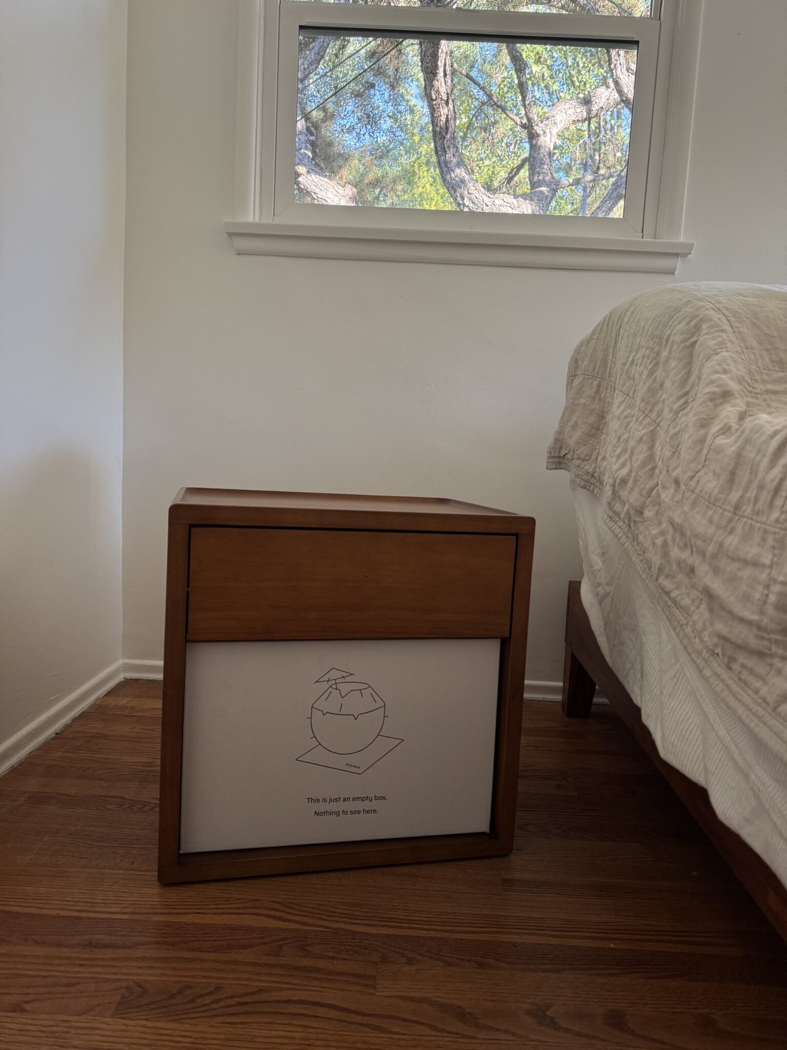 A wooden nightstand with one drawer sits next to a bed in a sunlit room. The front panel displays an illustration and text. A window with a tree view is above the nightstand.