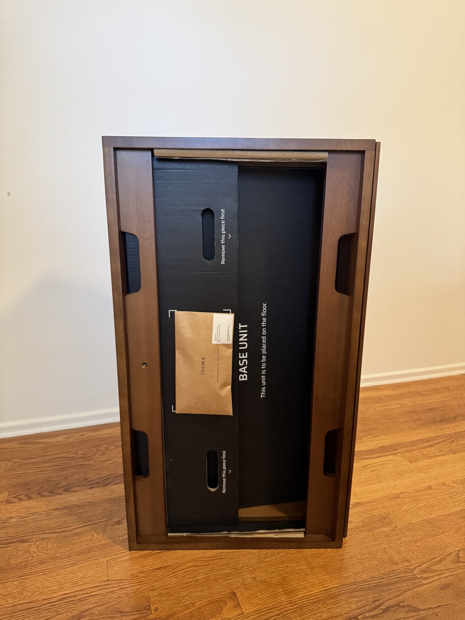 A wooden table is flipped upside down on a hardwood floor, showing packaging materials and a box labeled "BASE UNIT" inside the frame.