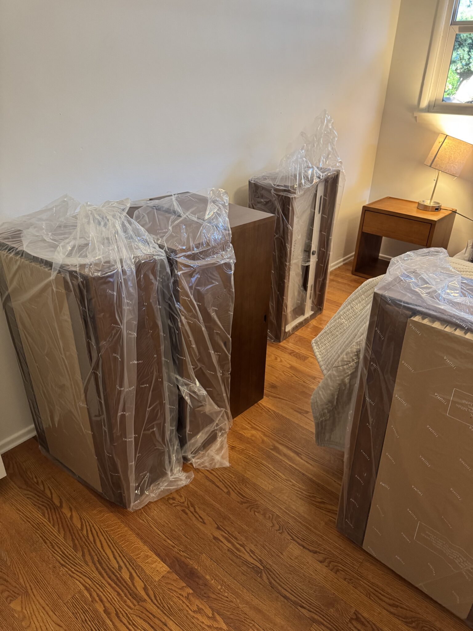 Several pieces of wooden furniture wrapped in protective plastic are placed along a bedroom wall, next to a window and a nightstand with a lamp.