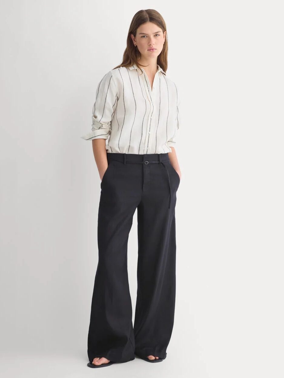 A person stands with hands in pockets, wearing a white button-up shirt with black vertical stripes and wide-leg black trousers against a plain background.