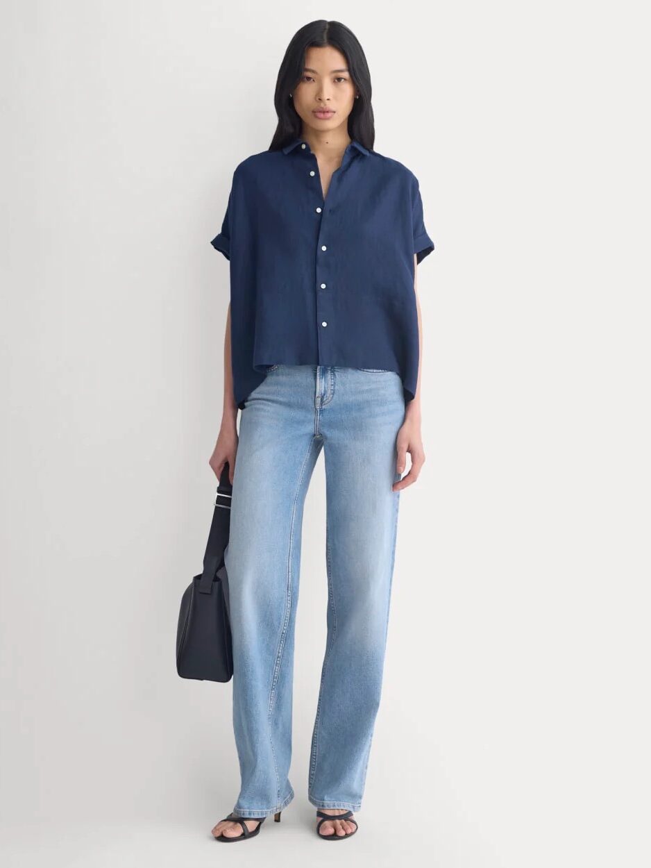 A person stands against a plain background wearing a navy short-sleeve button-up shirt, light blue jeans, black sandals, and holding a black bag.