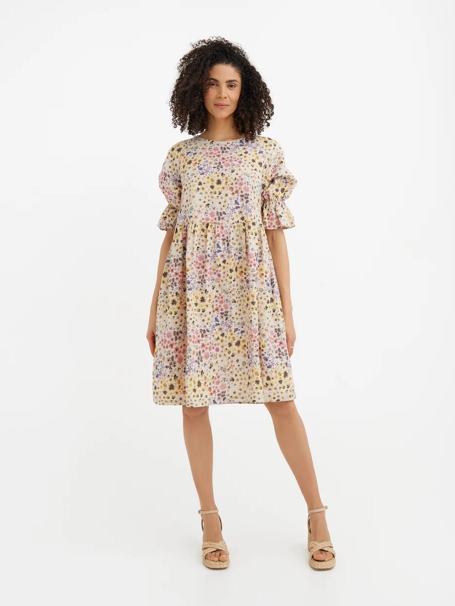 A woman stands facing forward wearing a knee-length, puff-sleeve dress with a multicolored floral print and beige platform sandals against a plain white background.