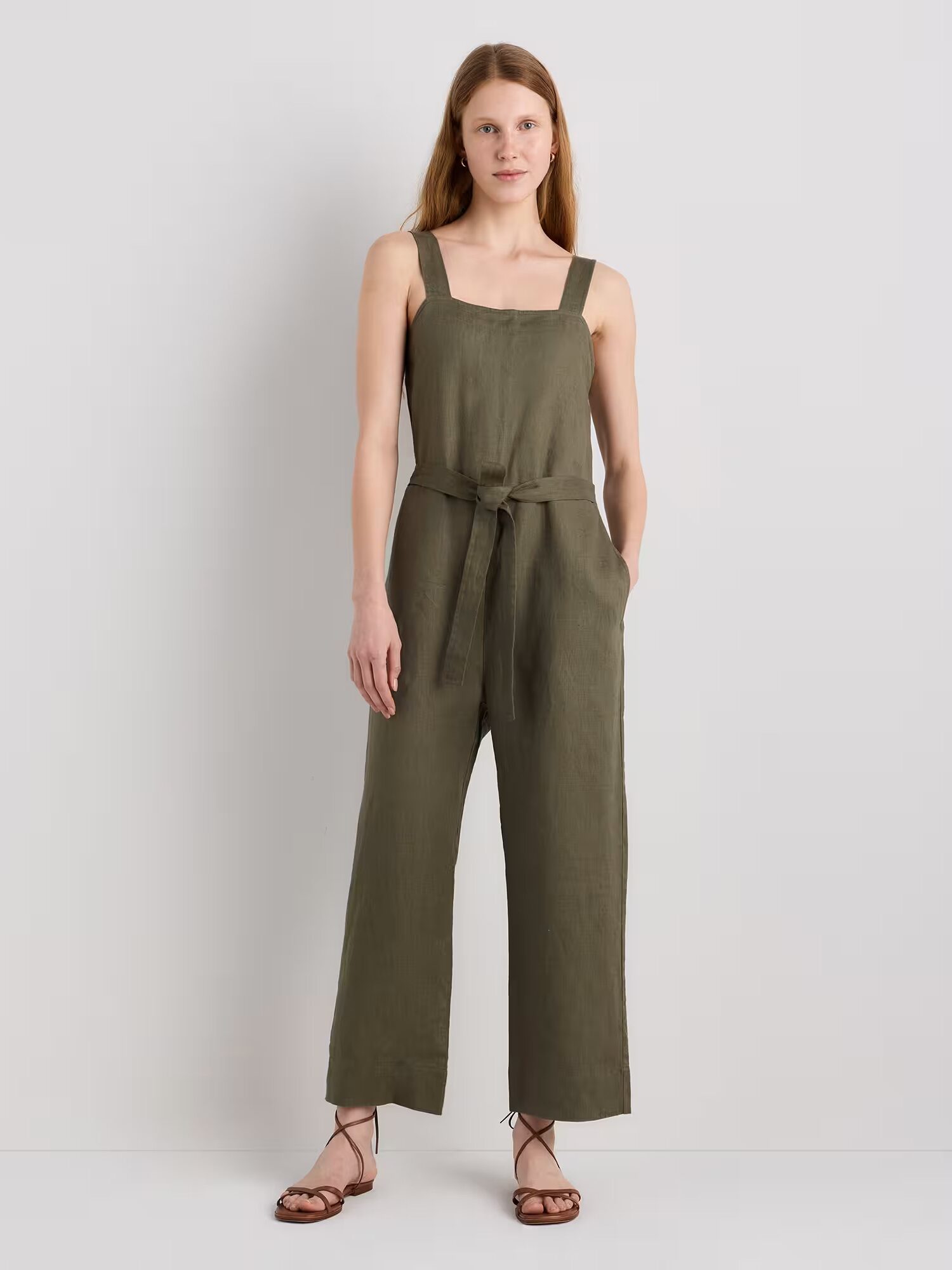 A woman stands against a plain background wearing an olive green sleeveless jumpsuit with a waist tie and wide legs, paired with brown flat sandals.