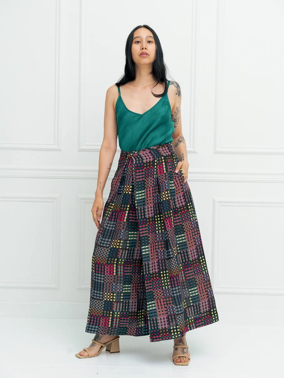 Woman standing against a white wall, wearing a green sleeveless top, a colorful patterned long skirt, and beige sandals.