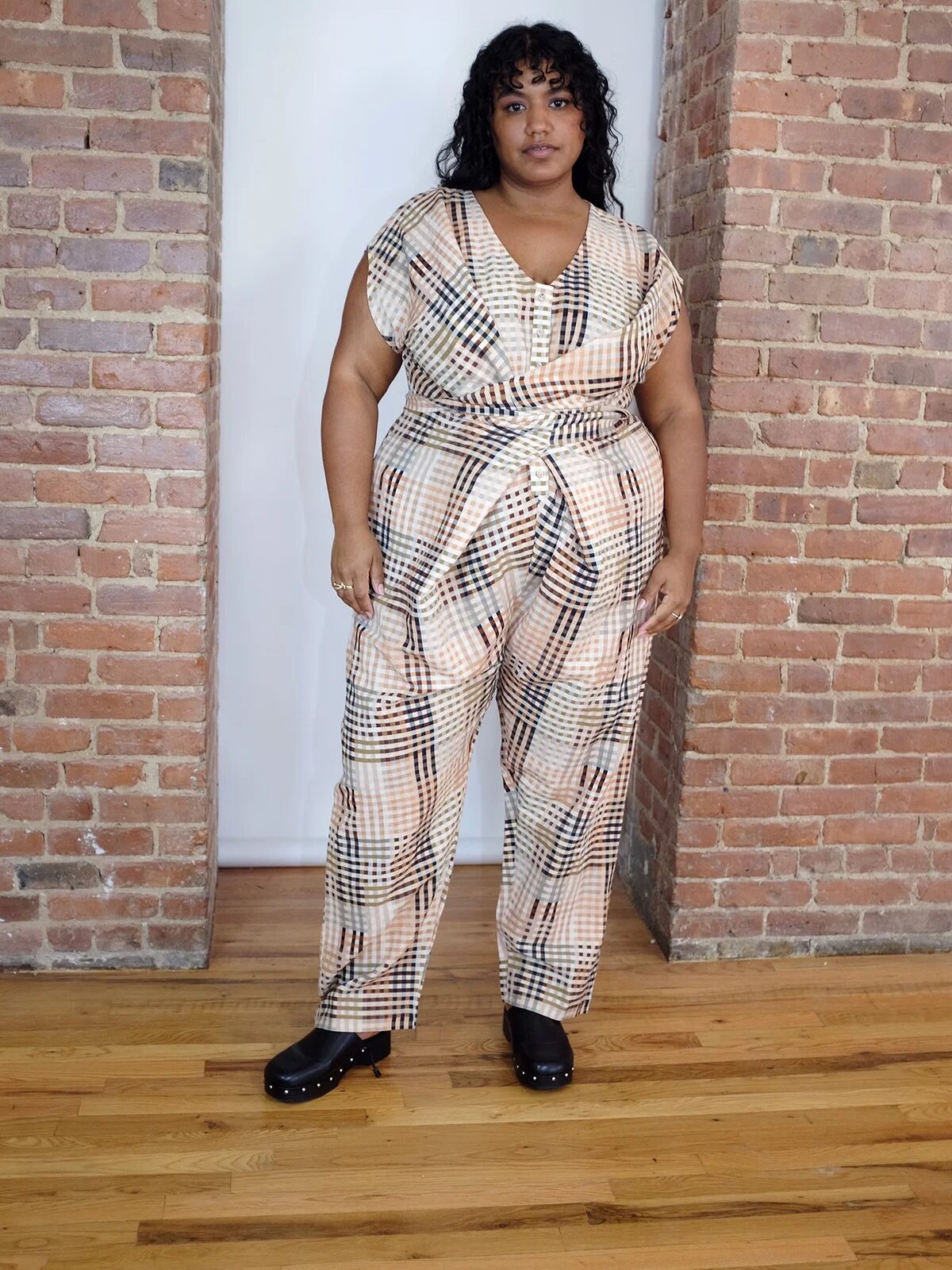 A person stands indoors against a brick wall, wearing a plaid jumpsuit and black clogs on a wooden floor.