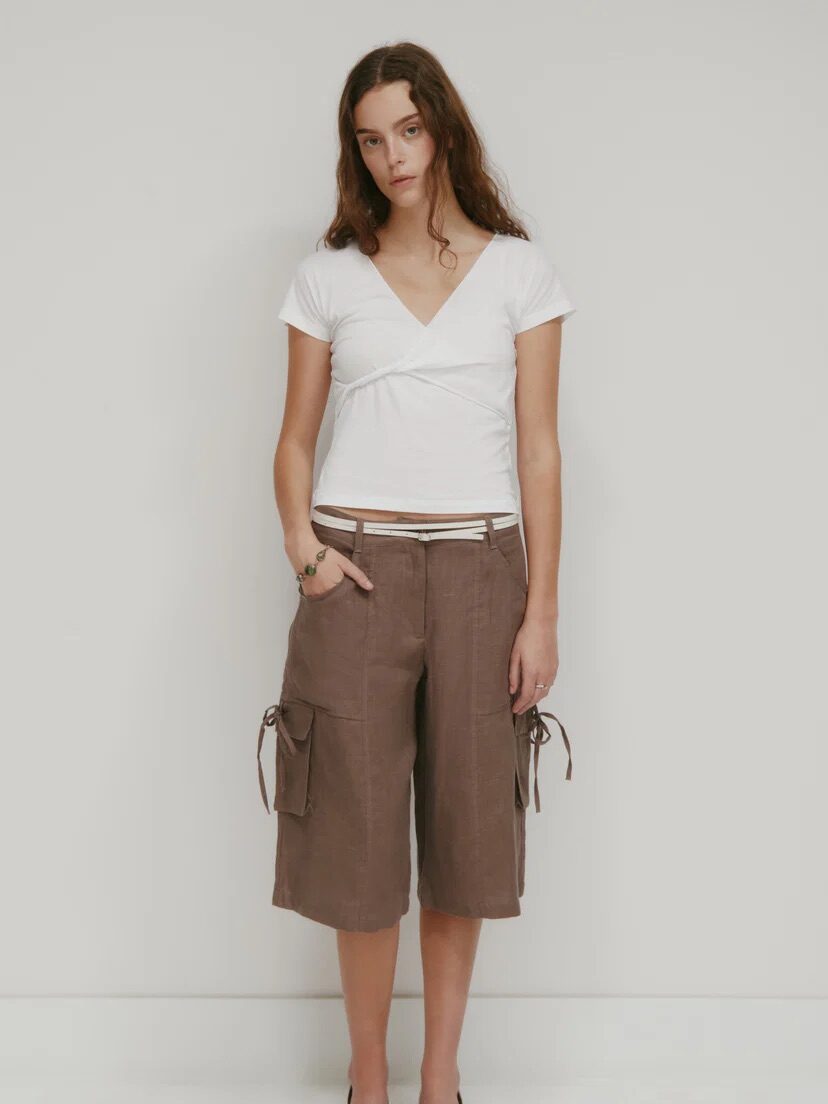 A woman stands against a plain background wearing a white V-neck short sleeve top, brown knee-length cargo shorts, a brown belt, and white flats.
