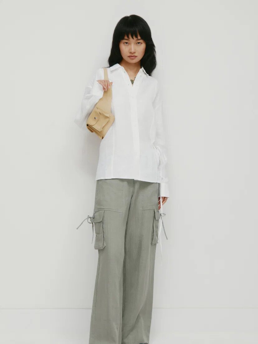 A person wearing a white button-up shirt, light gray wide-leg cargo pants, brown shoes, and holding a beige handbag stands against a plain white background.