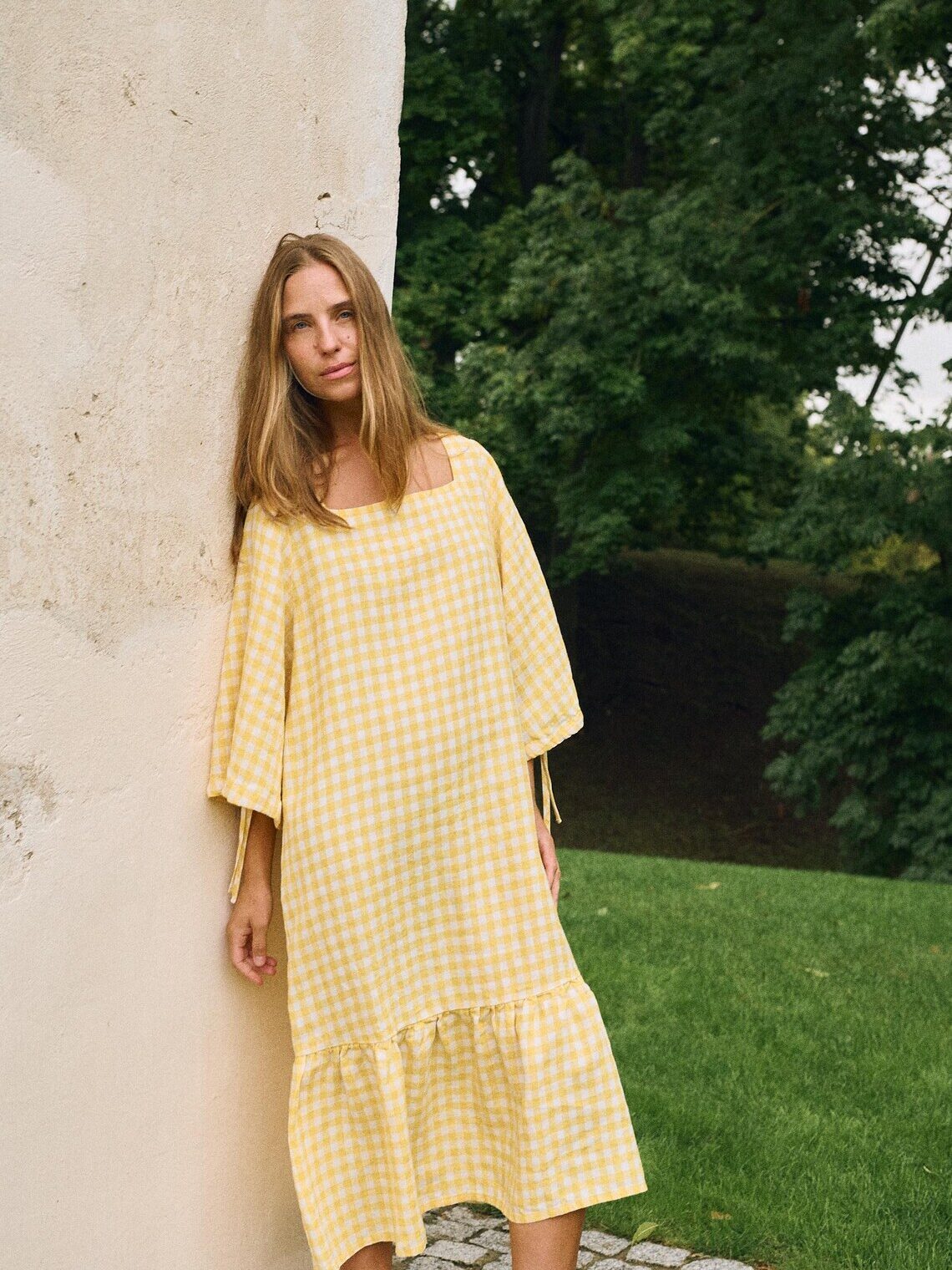 A woman wearing a loose-fitting yellow and white checkered dress stands against a light-colored wall, with green grass and trees in the background.