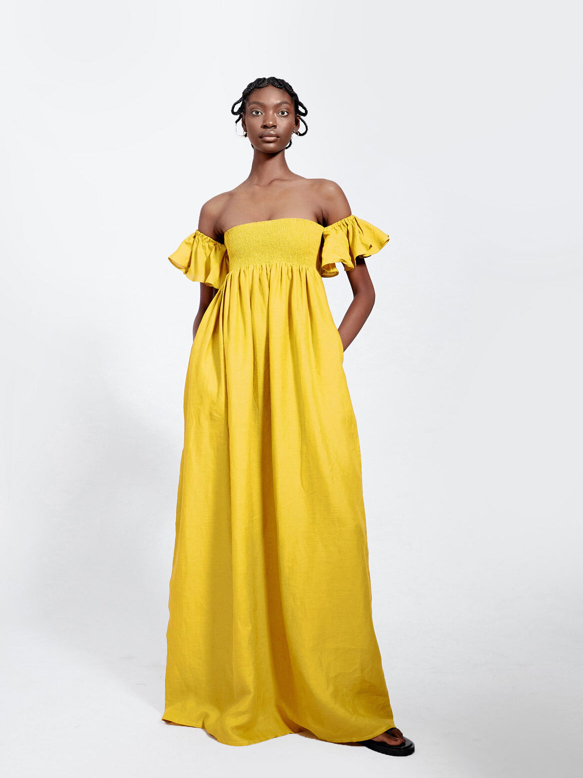 A person wearing a long, off-the-shoulder yellow dress with ruffled sleeves and black flat shoes stands against a plain white background.