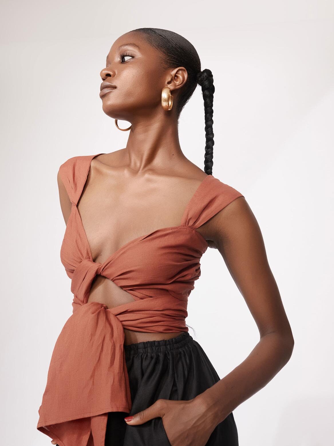Woman wearing a rust-colored knotted crop top and black pants, posing with one hand in her pocket against a plain background.