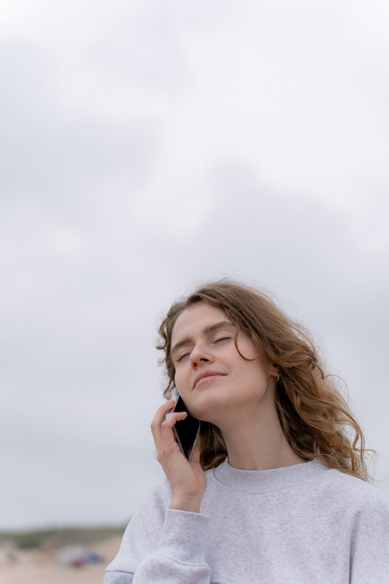 A woman with wavy hair holds a smartphone to her ear and stands outdoors under a cloudy sky, appearing calm with her eyes closed.