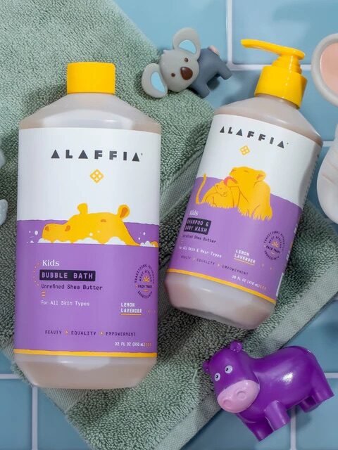 Two bottles of Alaffia Kids bubble bath with yellow and purple labels are placed on a green towel, surrounded by toy animals on a blue tiled surface.