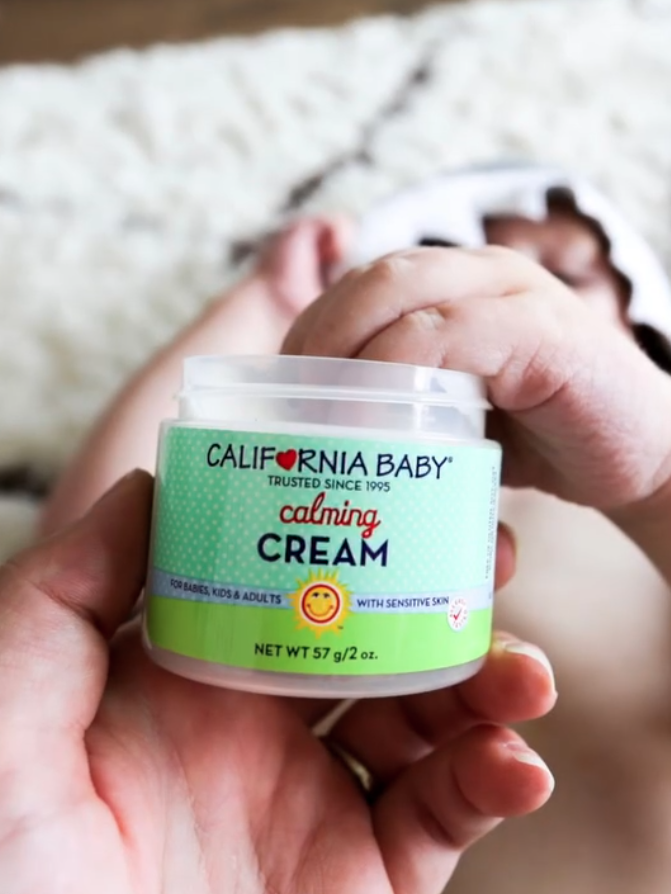A hand holds a container of California Baby calming cream in front of a baby lying on a soft, white rug.