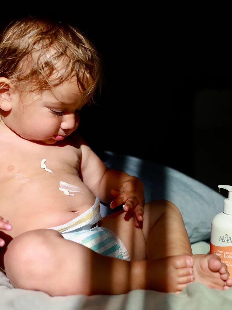 A baby wearing a diaper sits on a bed with lotion on its chest. A bottle of baby lotion is placed nearby.