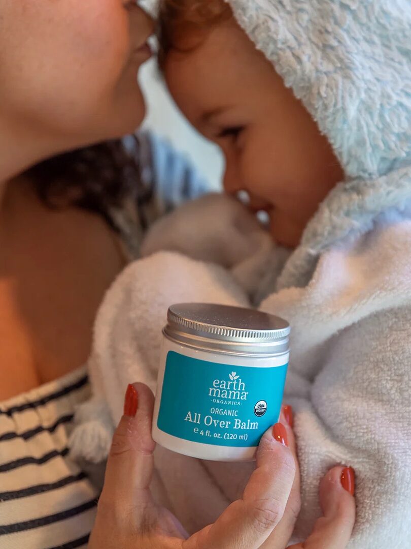 A woman holds a smiling baby in a hooded towel while displaying a jar of Earth Mama Organic All Over Balm.