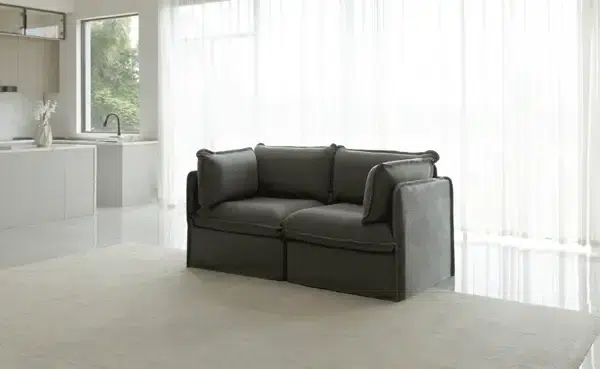A dark gray, cushioned loveseat sits on a light carpet in a modern, minimalistic living space with large windows and sheer curtains.